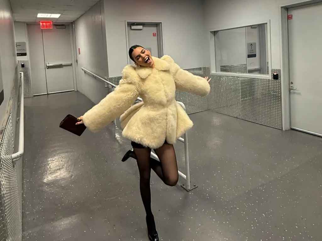 Kendall Jenner poses in a cream fur coat and black tights, laughing with her arms extended.