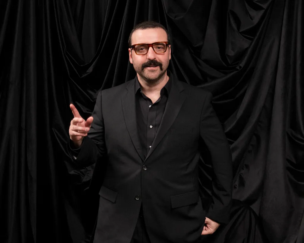 David Krumholtz in a black suit and glasses, pointing, in front of a black backdrop.