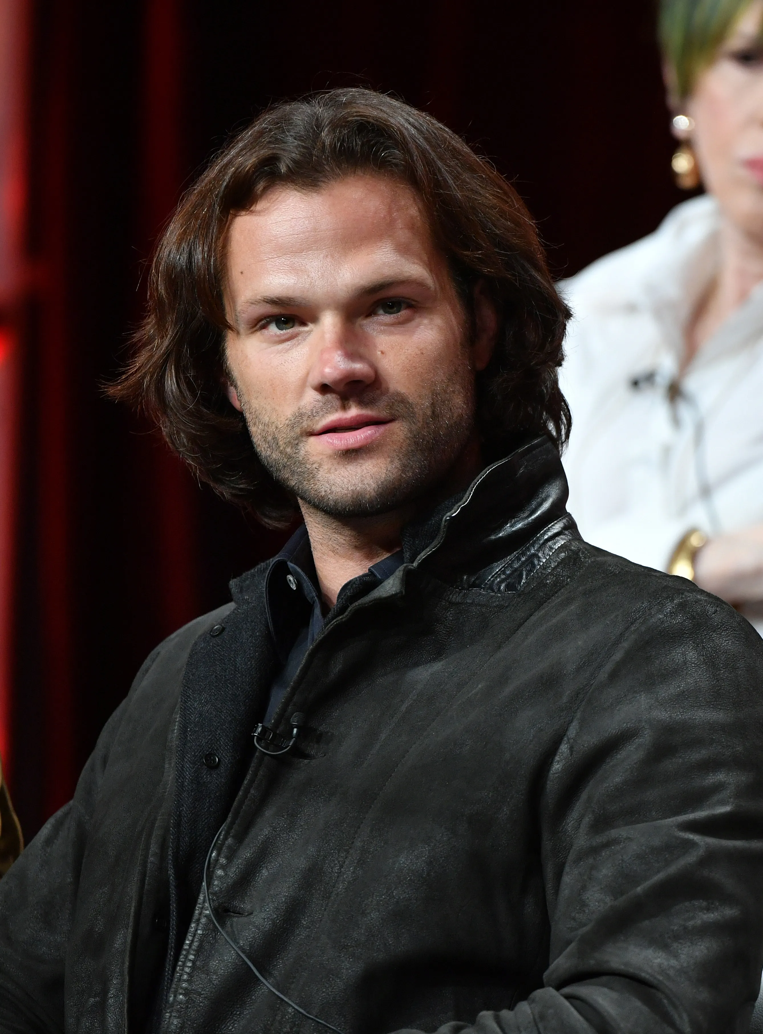 Jared Padalecki with long brown hair, a short beard, and a black jacket, looking at the viewer.