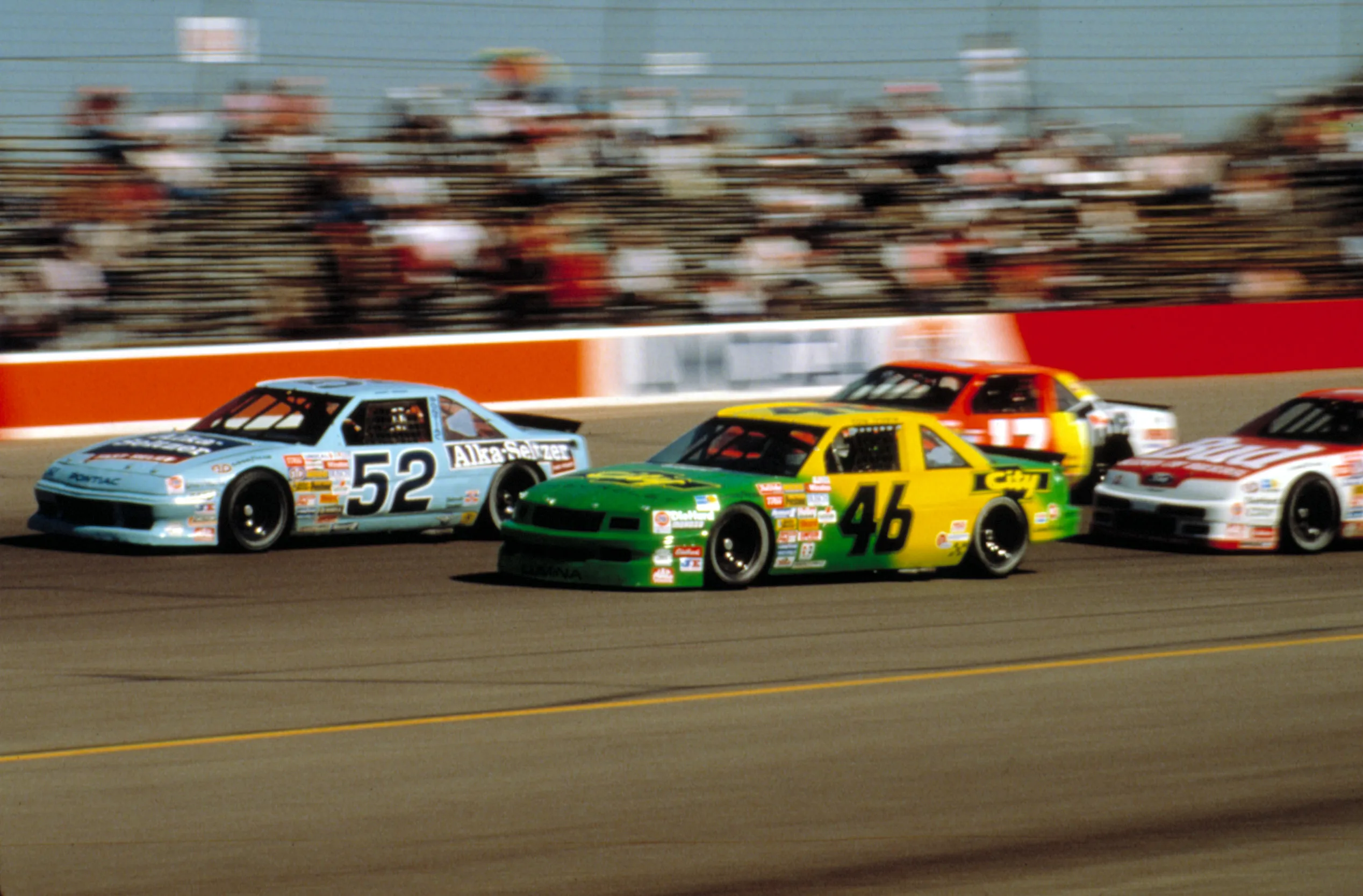Four race cars, including a light blue #52 and a green and yellow #46, speed around a track from the film 