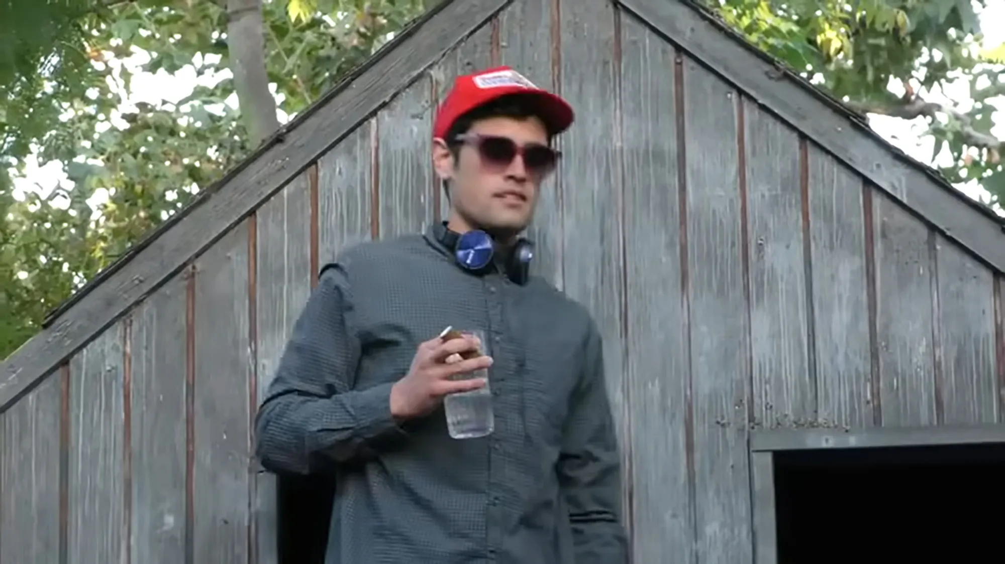 Nick Reiner wearing a red hat, sunglasses, and headphones around his neck, holding a glass.