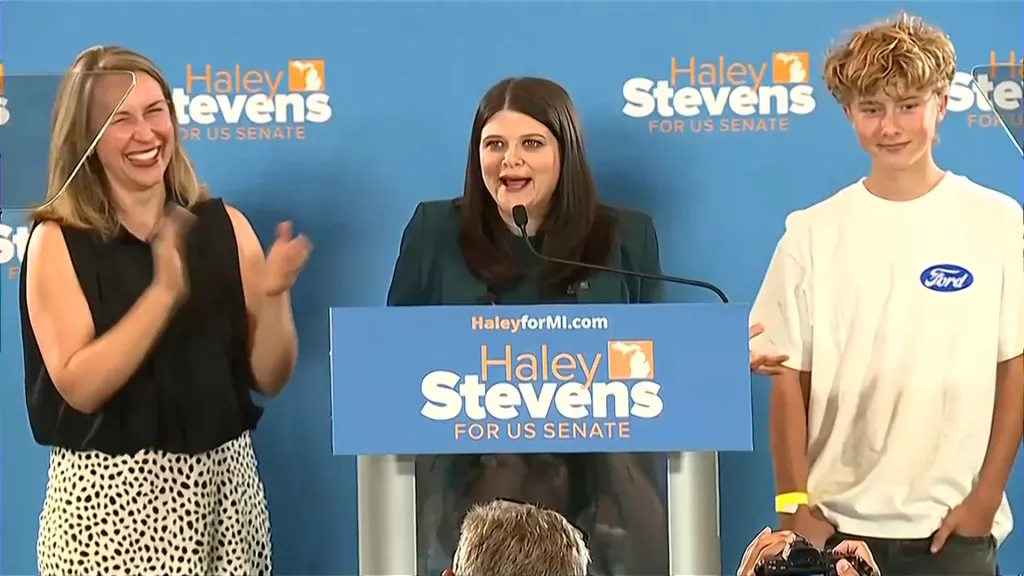 Democratic US Senate candidate Haley Stevens speaks at a podium while a supporter claps and a young man stands by, all against a blue backdrop with 