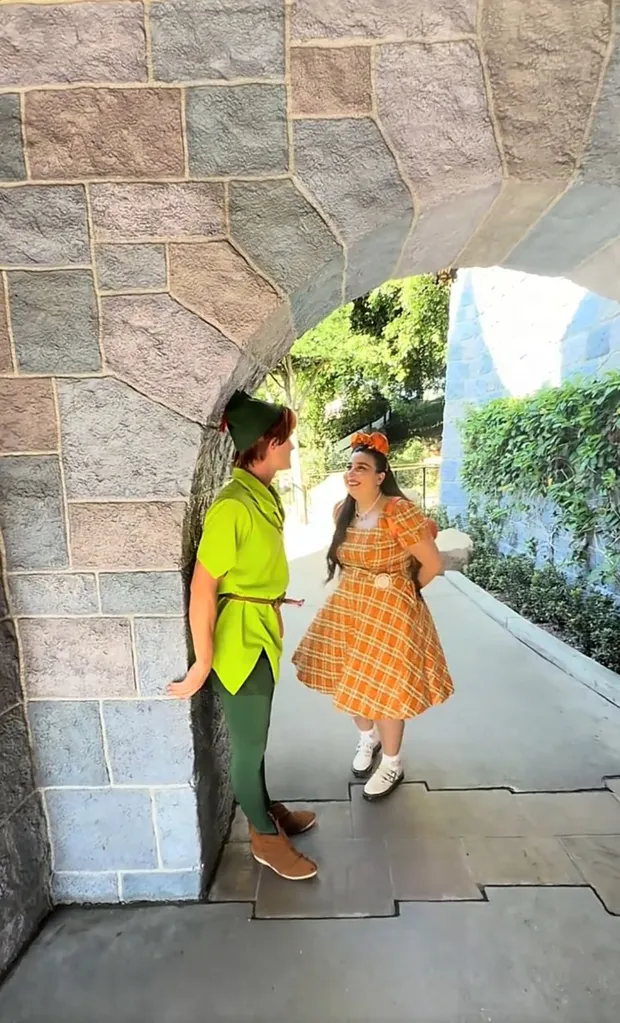 A young woman in an orange plaid dress and Peter Pan pose under a stone archway.