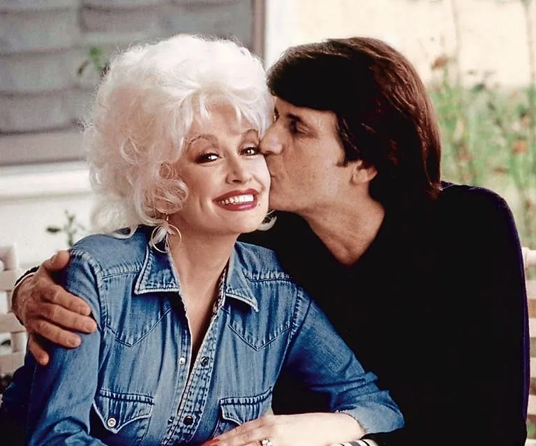 Dolly Parton smiling as her husband Carl Thomas Dean kisses her cheek.