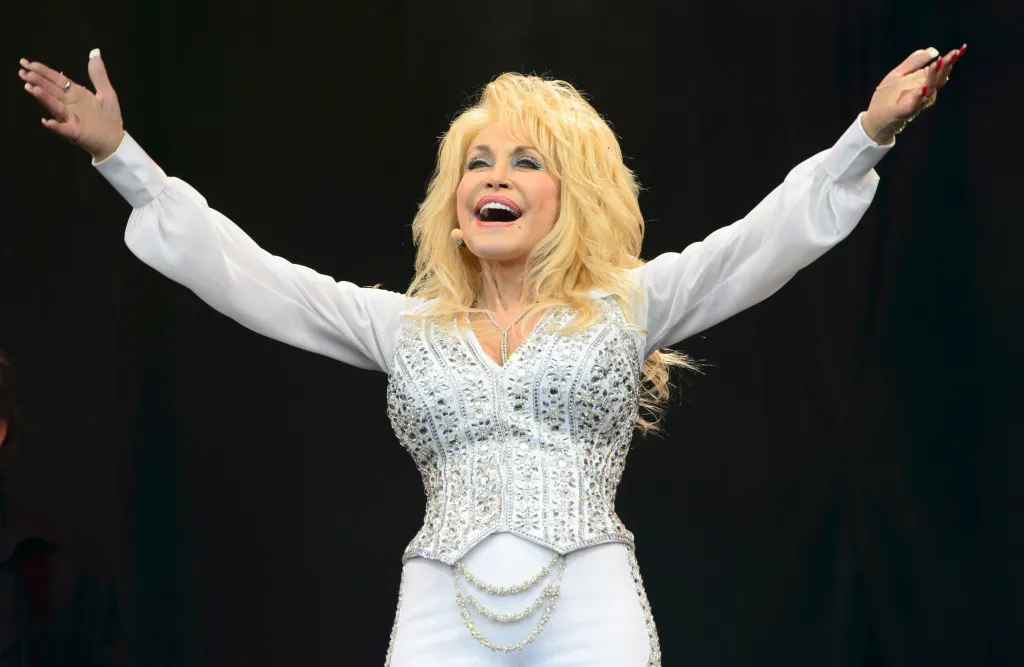 Dolly Parton singing with arms raised and mouth open, wearing a white bedazzled outfit.