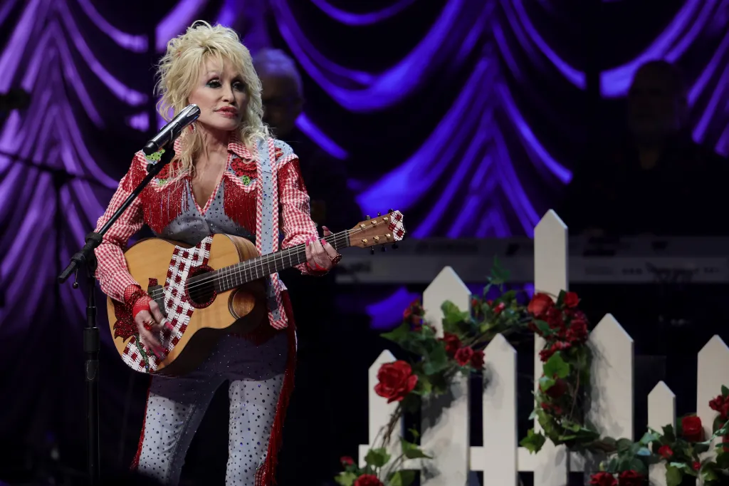 Dolly Parton performing on stage, wearing a red and white checkered jacket with red fringes, playing a guitar.