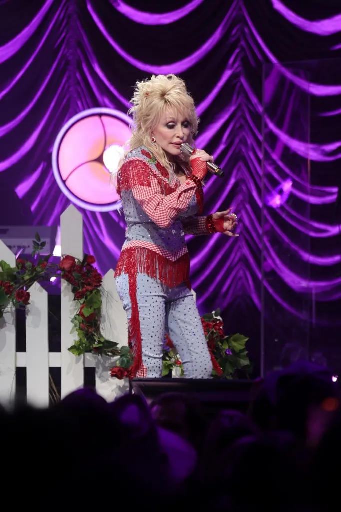 Dolly Parton performing on stage in a light blue and red outfit with fringe and polka dots, holding a microphone.