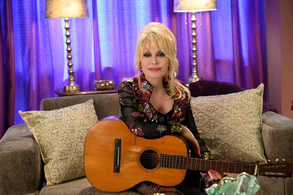 Dolly Parton holding a guitar while seated on a couch, looking at the camera.