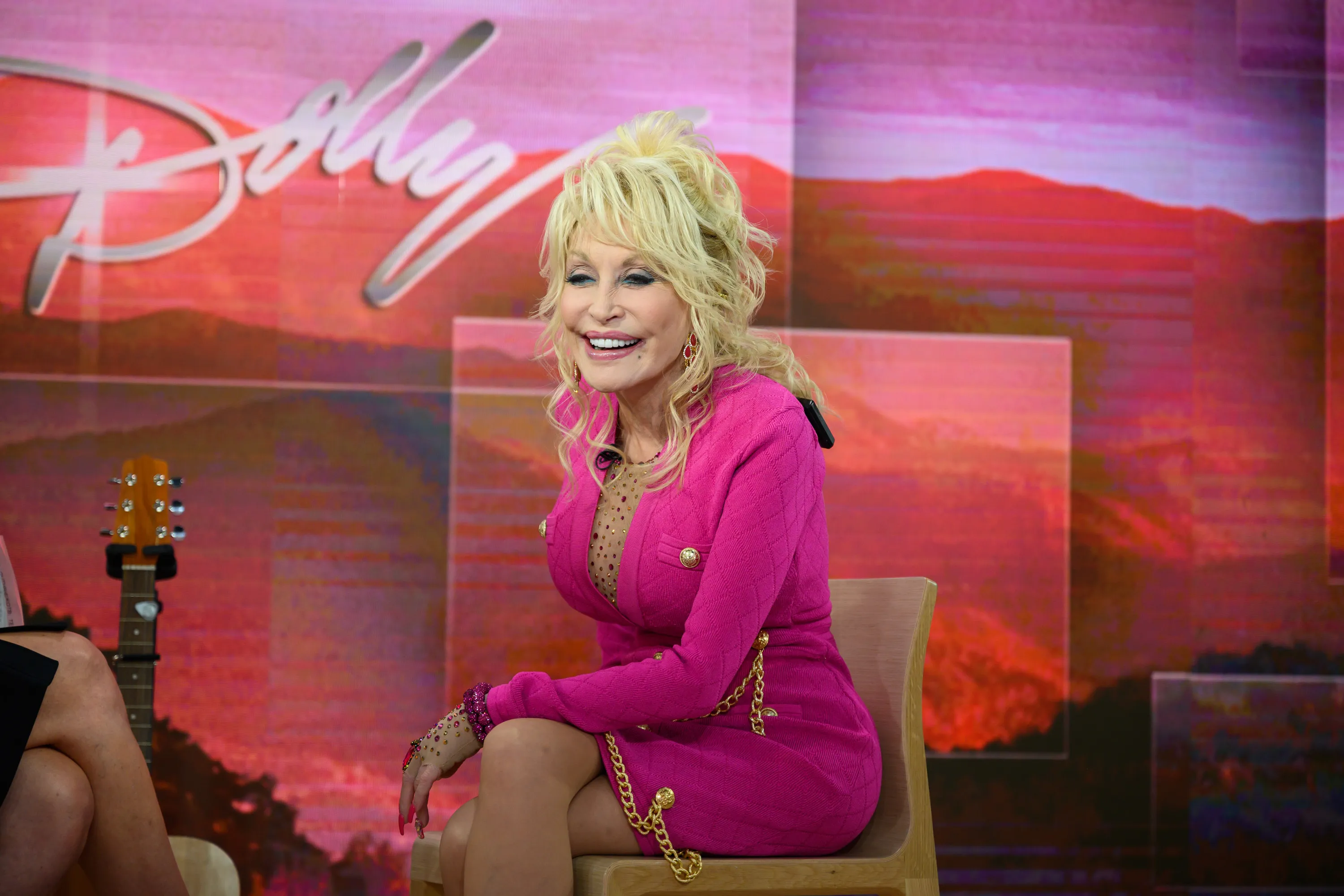 Dolly Parton smiles in a pink suit, with a guitar visible to her left, in front of a pink and red background with her name.