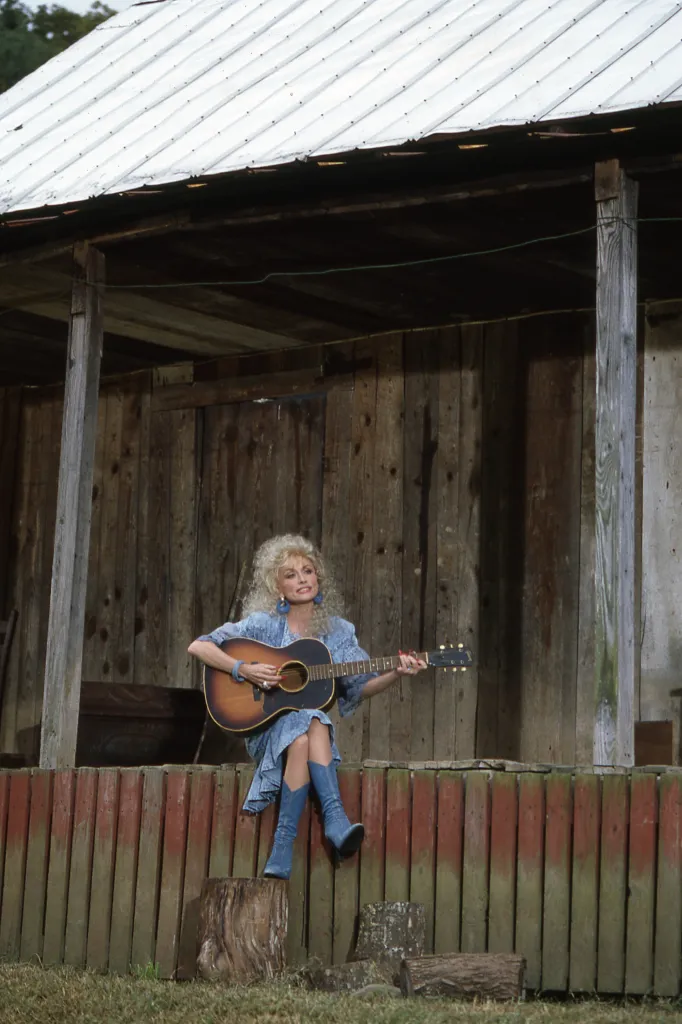 Dolly Parton in a denim dress and boots, seated on a red and green fence and playing a guitar in front of a wooden building with a metal roof.