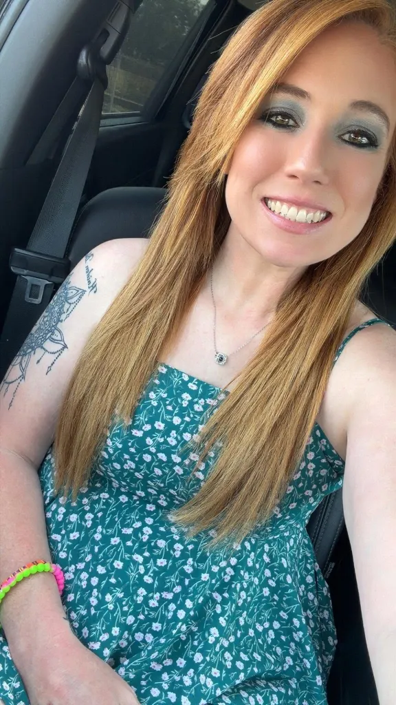 Christen Phillips smiling in a car, wearing a green floral dress and a seatbelt.