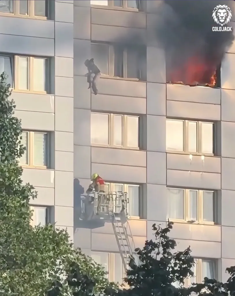 A woman dangling from an 11th-floor window of a burning building prepares to jump into a firefighter's basket on a ladder extended to the 9th floor.