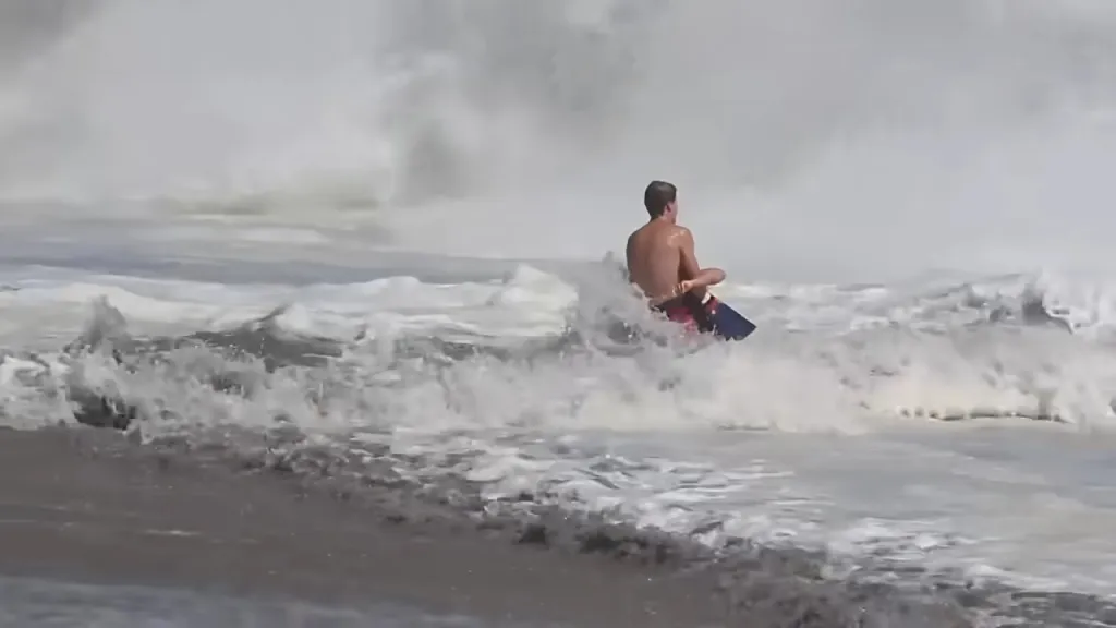 A lifeguard, shirtless and wearing swim fins, in rough waves with a large wave breaking behind him.
