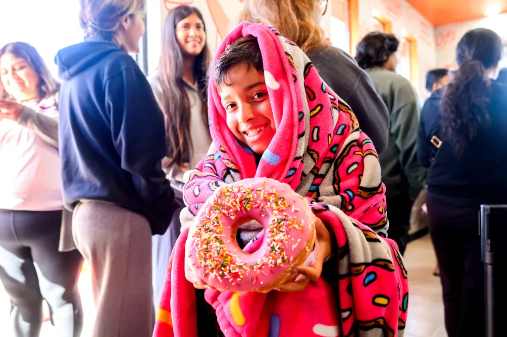 A young boy smiles and holds a large pink donut with sprinkles at Randy's Donuts grand opening.