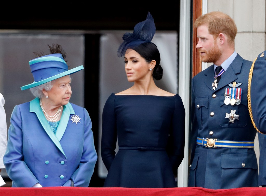 Queen Elizabeth II, in a blue coat with turquoise trim and matching hat, Meghan, Duchess of Sussex, in a navy blue dress and blue hat, and Prince Harry, Duke of Sussex, in a blue military jacket and medals, watch a flypast from a balcony.