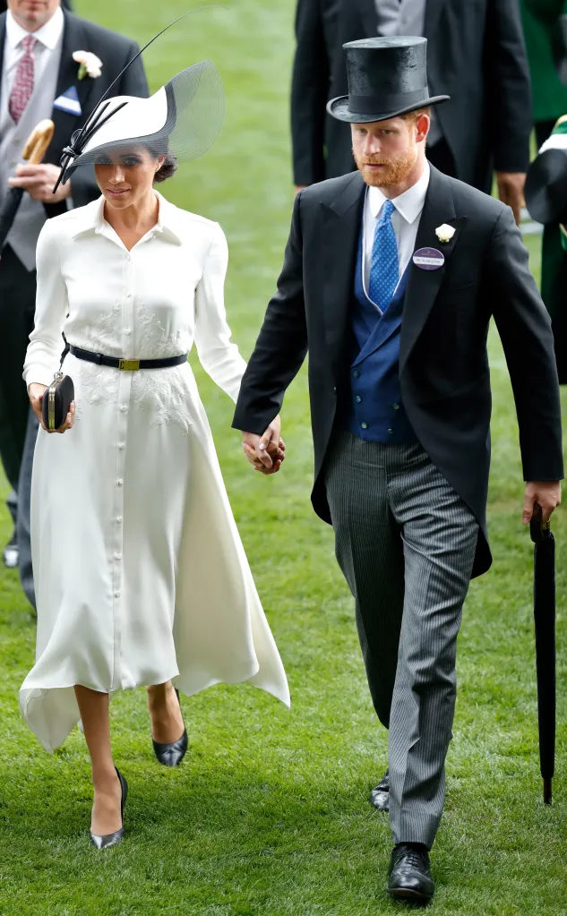Meghan, Duchess of Sussex, in a white shirt dress with black belt and white hat, and Prince Harry, Duke of Sussex, in a morning suit and top hat, walk hand-in-hand across a grassy field at Royal Ascot.