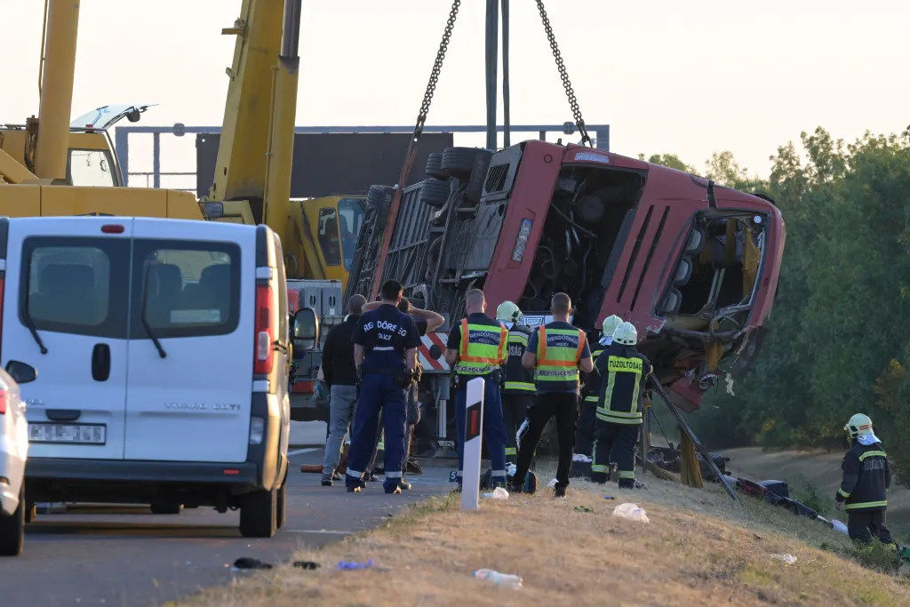Emergency services attending the scene of a tourist bus accident in Mezokeresztes, Hungary, showing a damaged red bus being lifted by a crane.