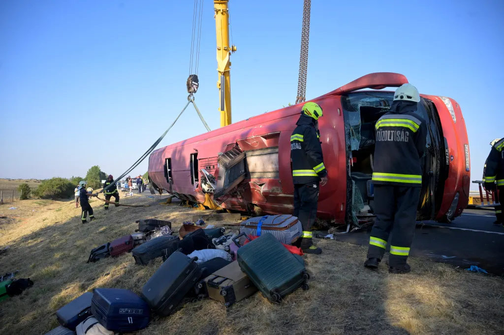 Emergency services attend to an overturned red tourist bus in a ditch, with scattered luggage and a crane attempting to upright the vehicle.