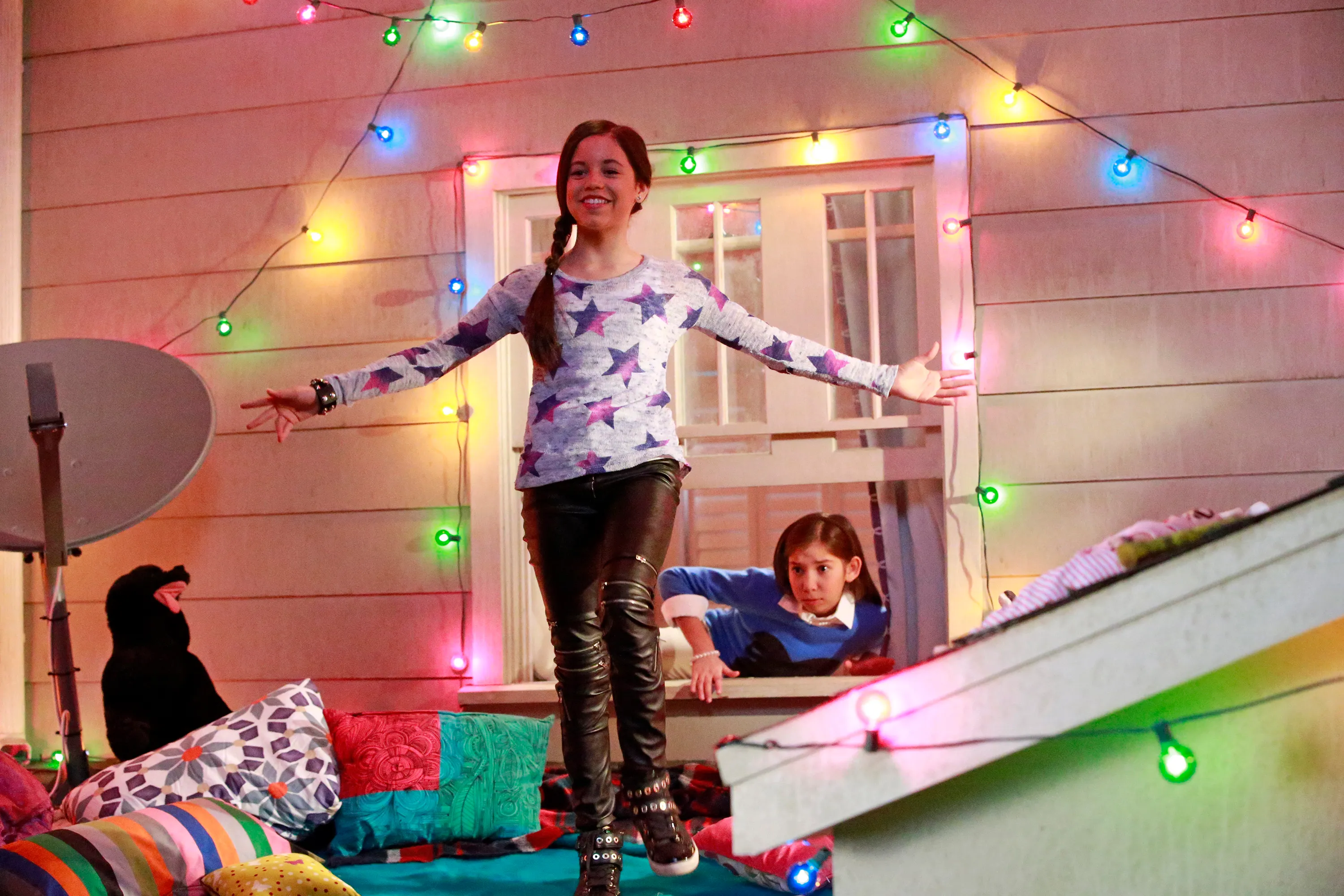 Two girls posing, one standing with arms out, another peeking out of a window, surrounded by colorful string lights.
