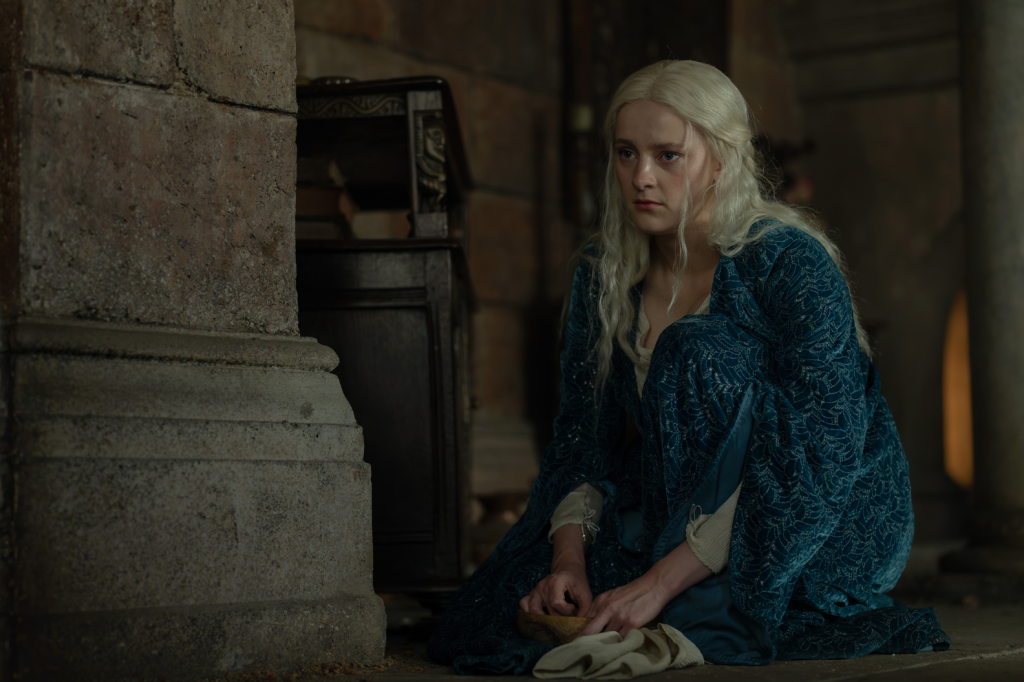 Phia Saban as Princess Helaena Targaryen in a blue robe, kneeling on the floor.