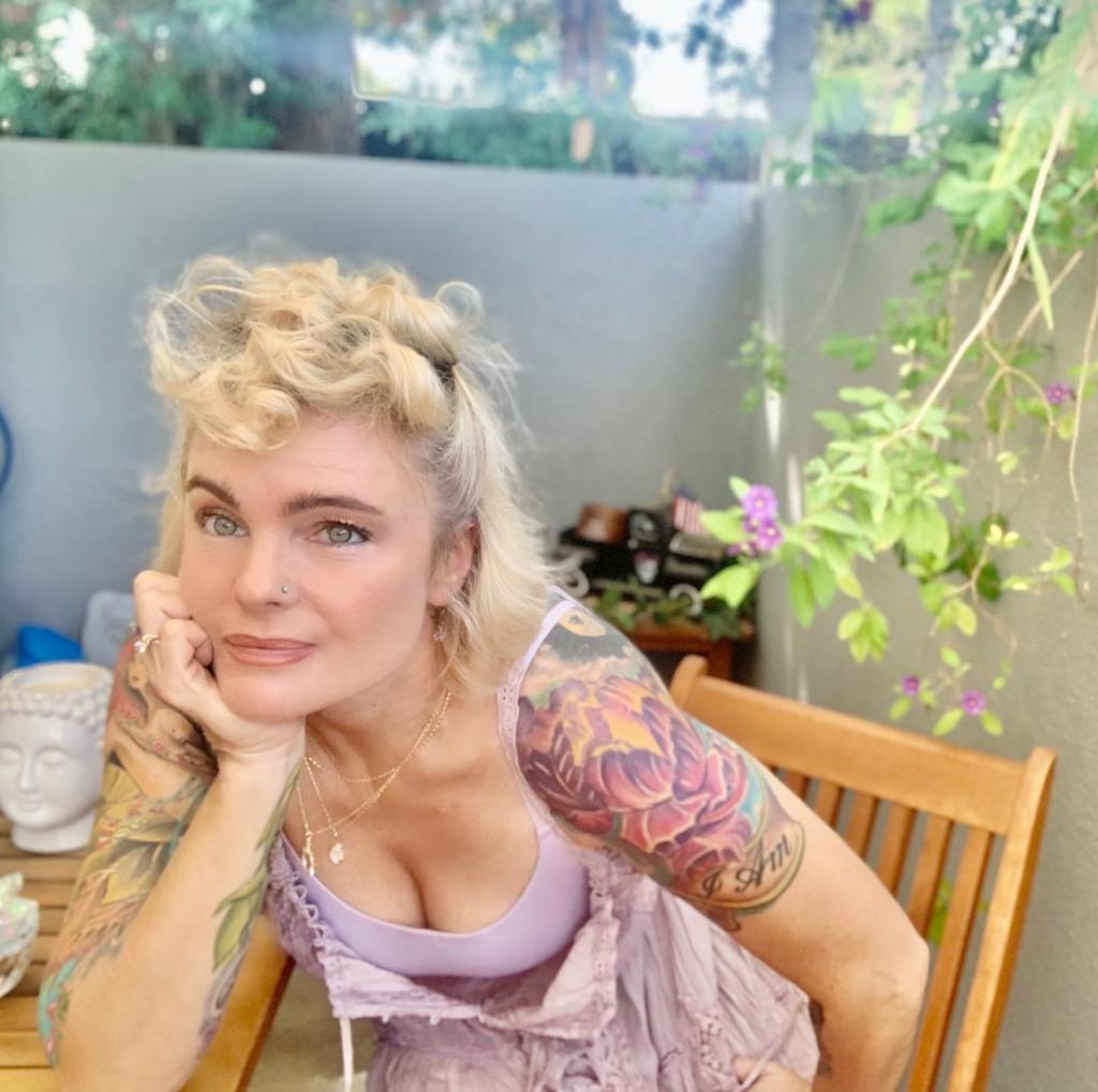A woman with blonde hair, tattooed arms, and a nose piercing, wearing a light purple top, leans on a wooden table outdoors.