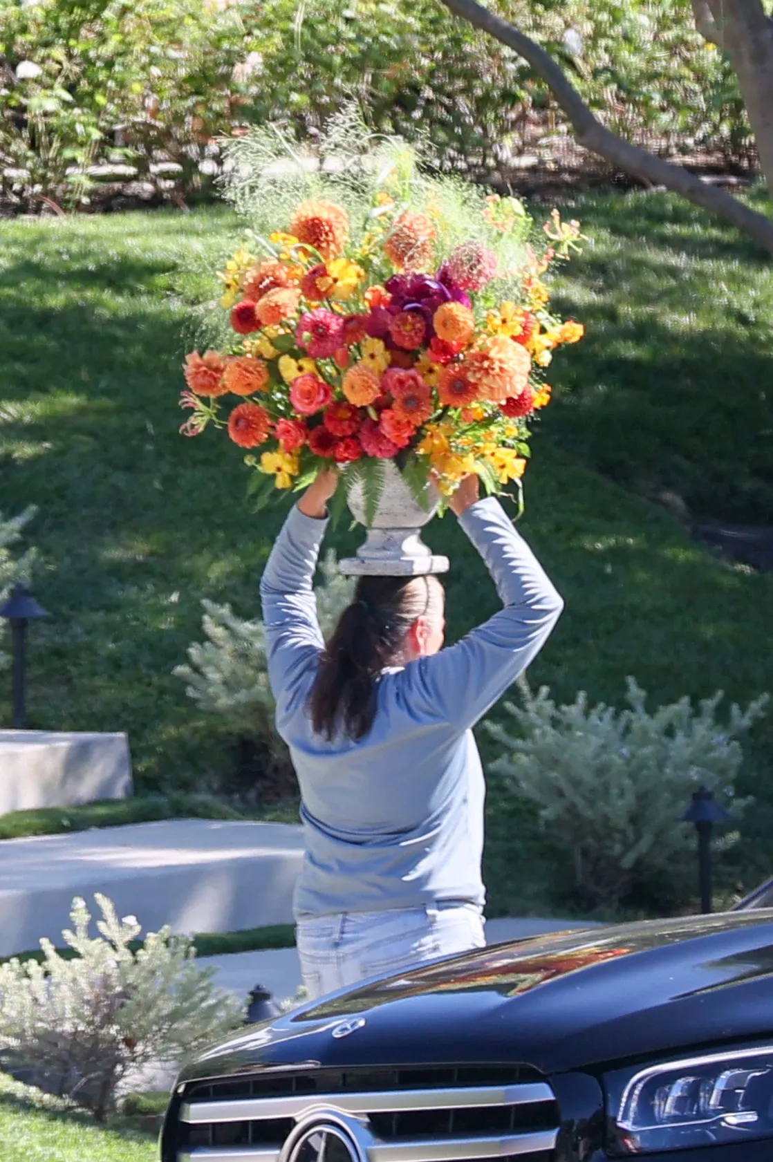 A person in a blue shirt carrying a large vase of colorful flowers on their head.