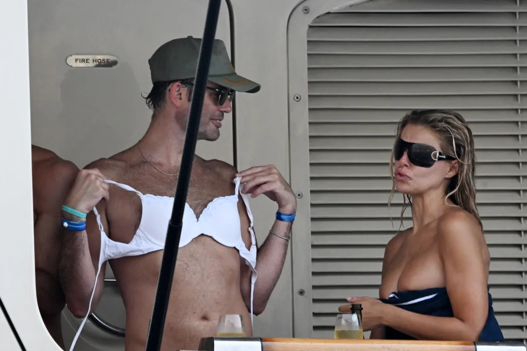 Jack Lumsden wears a white bra over his chest while Brooks Nader poses in a towel and sunglasses on a yacht.