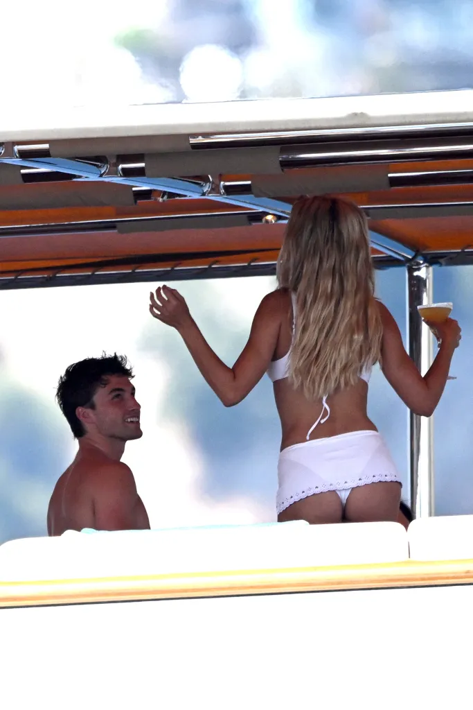 Brooks Nader in a white bikini on a yacht, holding a drink and looking away, with Jack Lumsden smiling beside her.