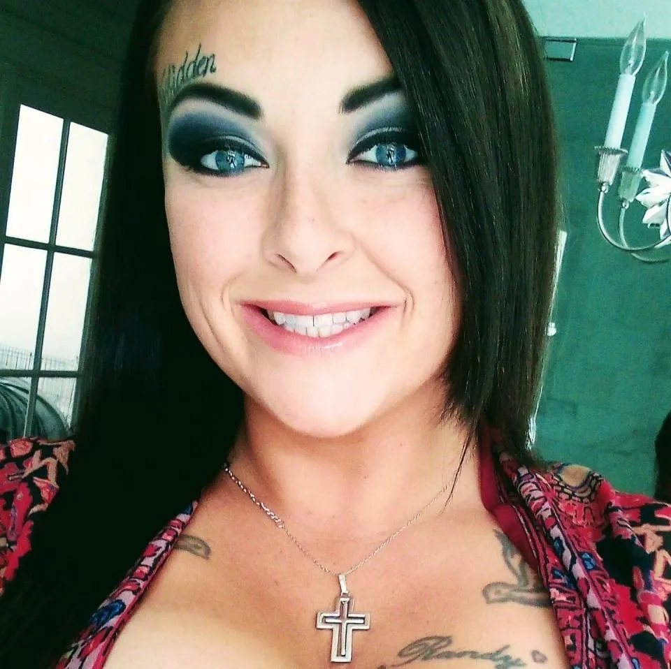 Grace Pauline Kelley smiles, wearing a cross necklace and a top with a colorful print, with a tattoo on her forehead that says 
