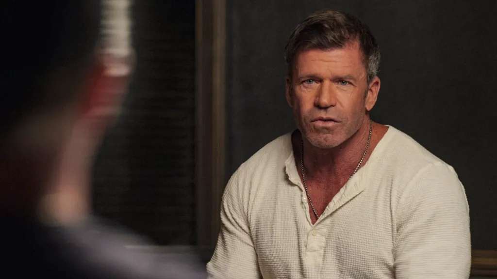 Taylor Sheridan, dressed in a white shirt and chain, looks intently at an unseen person.