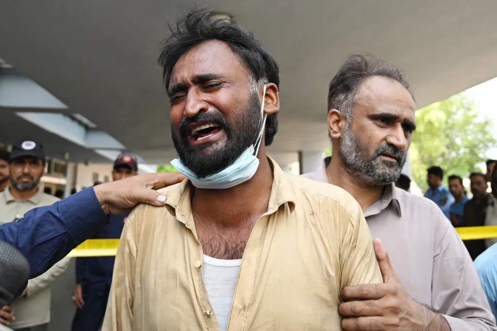 A man cries in anguish, his mouth open and eyes closed, while another man places a comforting hand on his shoulder following a fatal hospital fire.