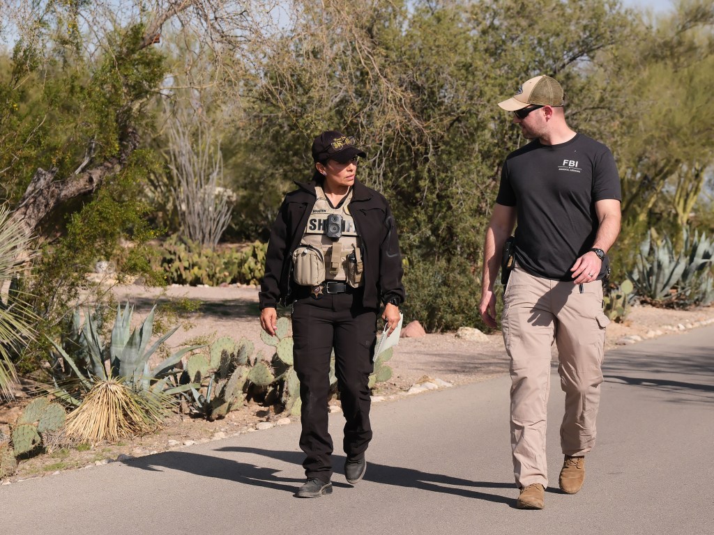 A female sheriff , in a black top and black pants nad black baseball cap with a khaki vest, and male FBI agent, in khaki pants, black T-shirt and khaki baseball cap, walk along a paved path in a desert landscape.