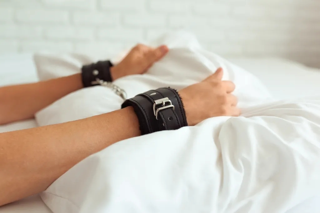 Female hands in black leather cuffs with a chain between them, lying on white bedding.