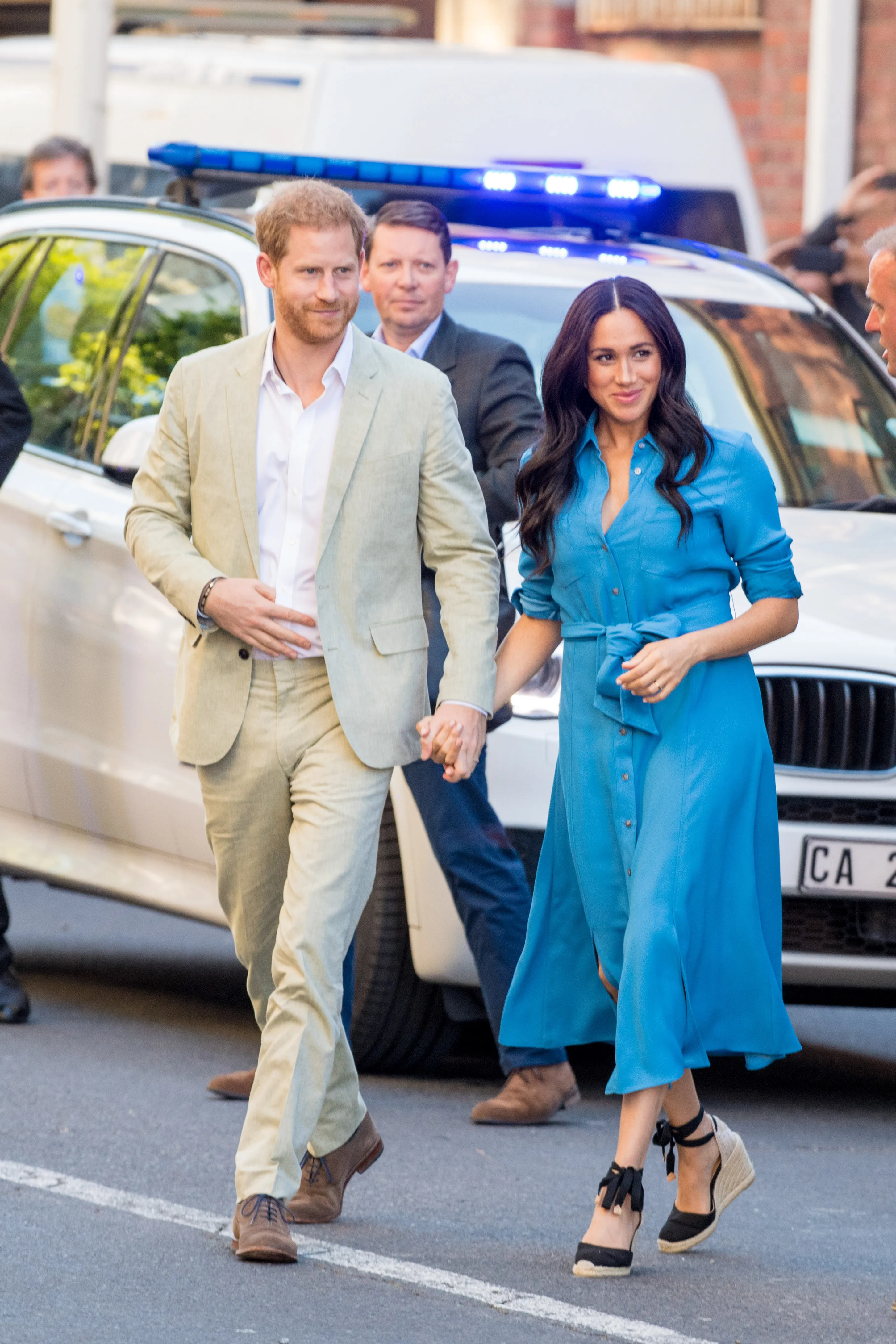 Meghan Markle in a blue dress and Prince Harry in a beige suit hold hands while walking with security.