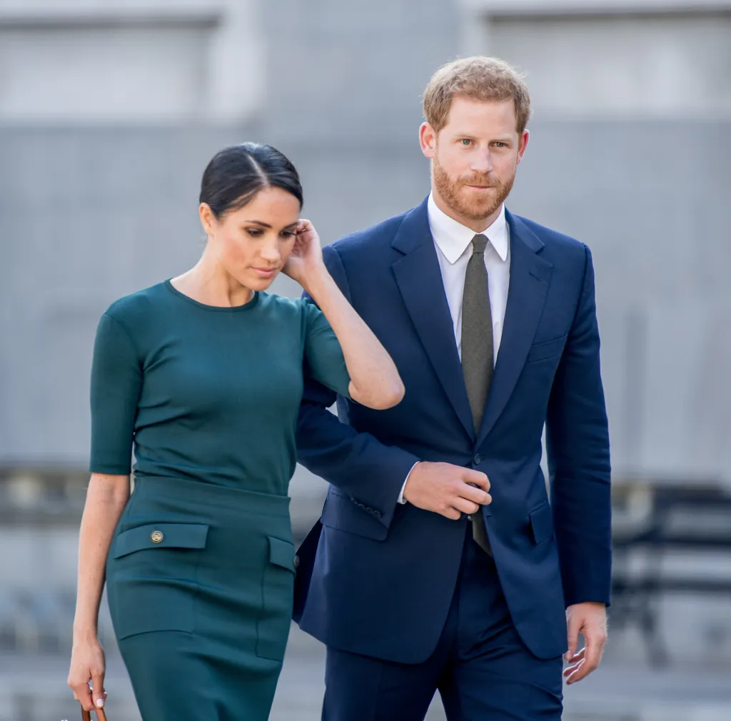 Meghan Markle in a green dress with her hand on her head, with Prince Harry in a blue suit next to her, looking forward.