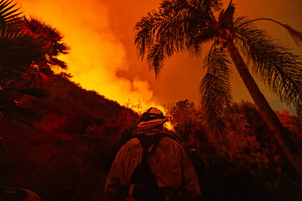 A firefighter stands guard in front of a hillside engulfed in orange flames and smoke.