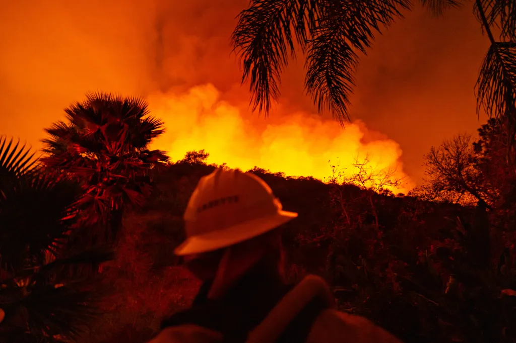 A firefighter in a hardhat stands guard as a wildfire glows brightly over a ridge behind them.