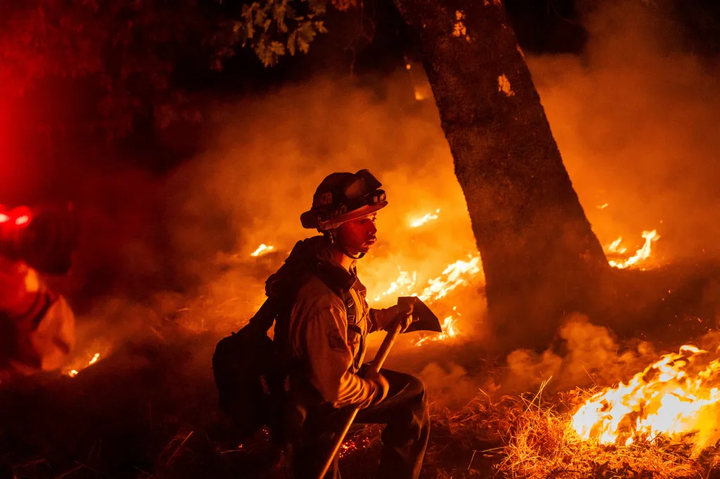 A firefighter in a helmet and protective gear, holding an axe, battles the Pickett Fire in Napa County, California, with flames visible around him.