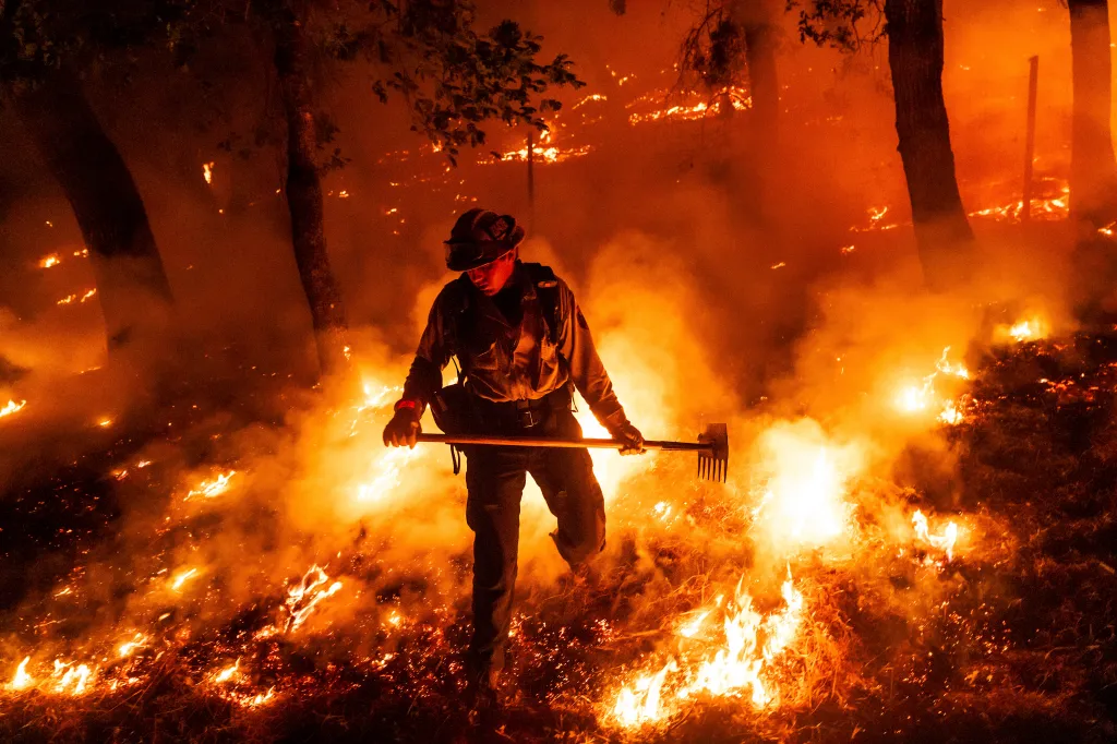 A firefighter battling the Pickett Fire in Napa County, California, holding a rake-like tool surrounded by flames.