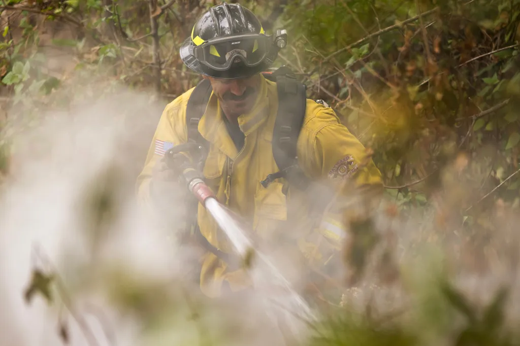 A firefighter in a yellow jacket and helmet sprays water on a hotspot.