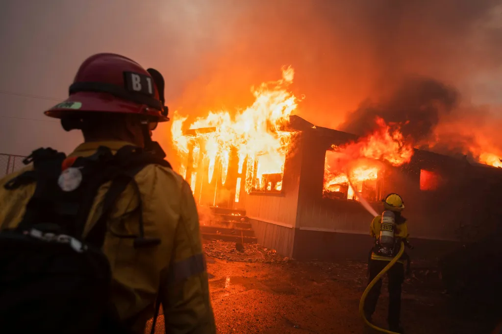 Firefighters battle the Palisades Fire in Los Angeles, with one directing a hose at a burning house and another looking on.