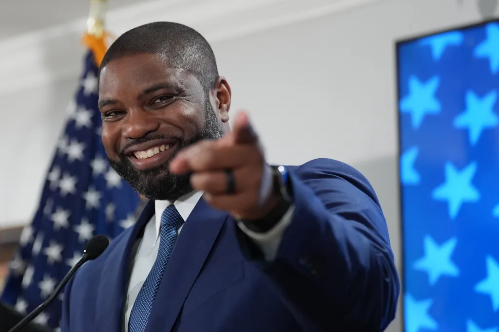 Byron Donalds, smiling, points forward while standing at a podium.