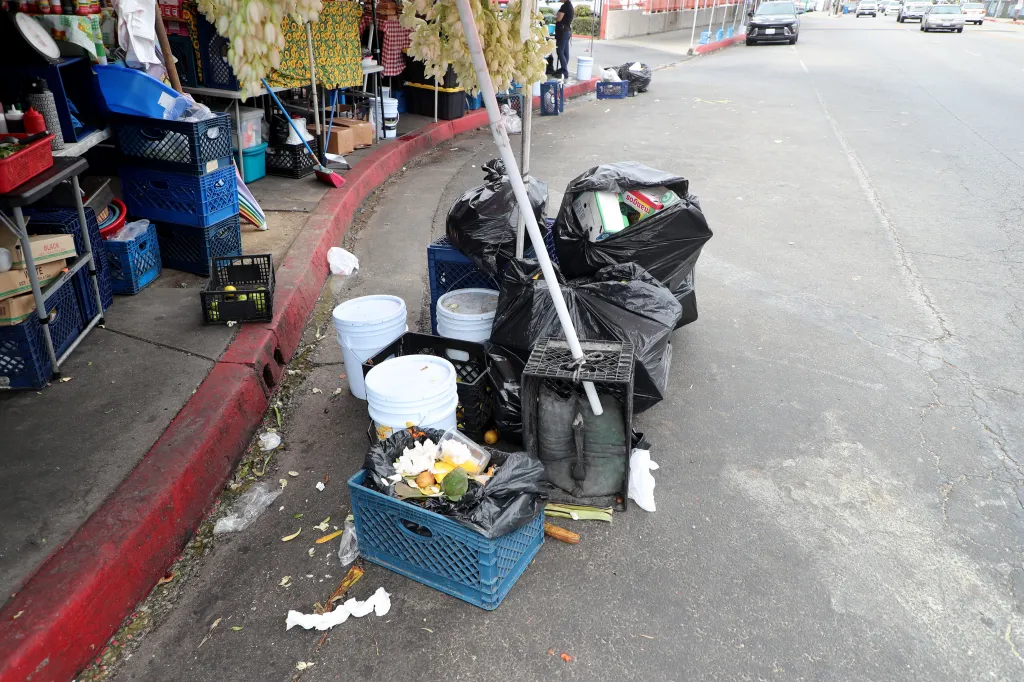 Food vendors on the Salvador Corridor in Los Angeles, California, with bins and black garbage bags on the sidewalk.