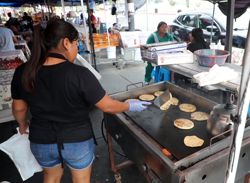 A food vendor flipping pupusas on a griddle in an outdoor market.