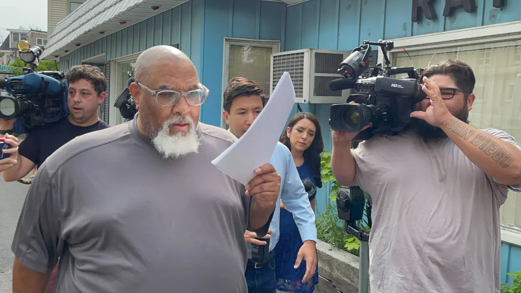 Cedric Lodge shielding his face with an indictment printout as he walks from the courthouse surrounded by reporters and cameramen.