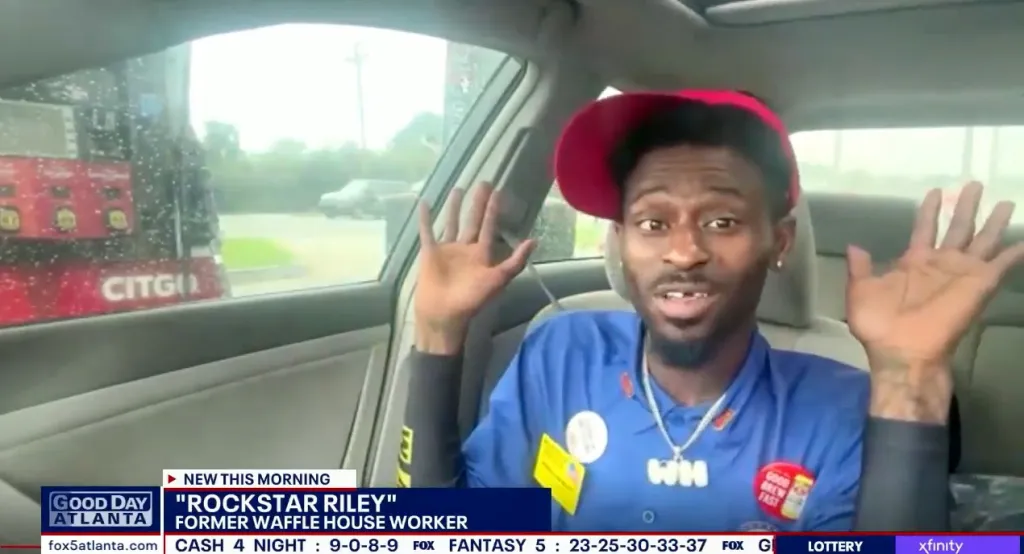 Rockstar Riley, former Waffle House employee, wearing his uniform and a red hat, raises his hands in a car.