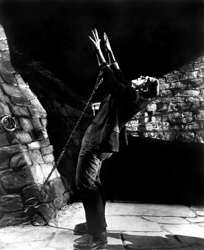 Boris Karloff as Frankenstein's Monster, chained in a dungeon with arms raised.