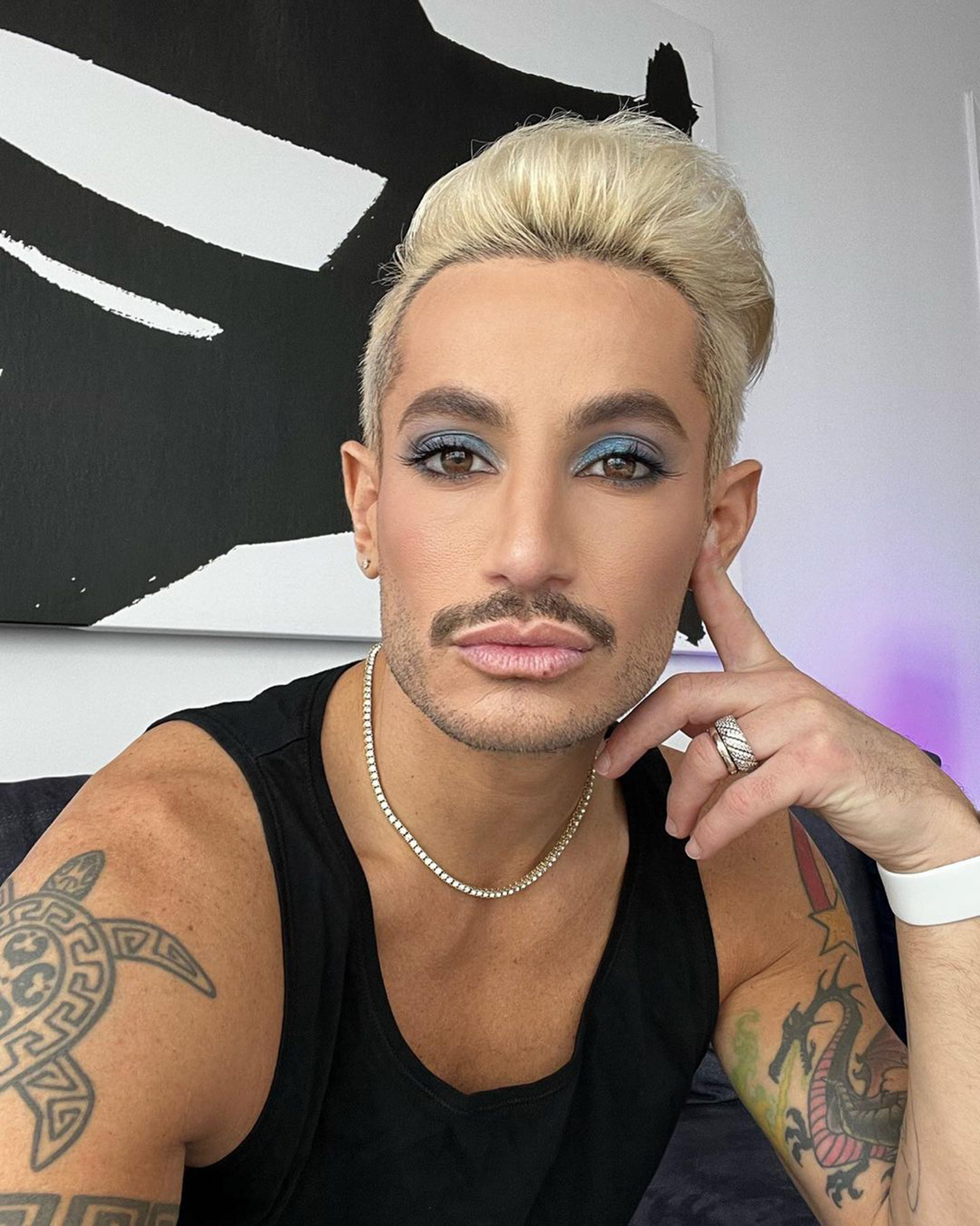 Frankie Grande looking at the camera, with a full face of makeup, including blue eyeshadow, with blonde hair, wearing a black tank top and a diamond necklace.