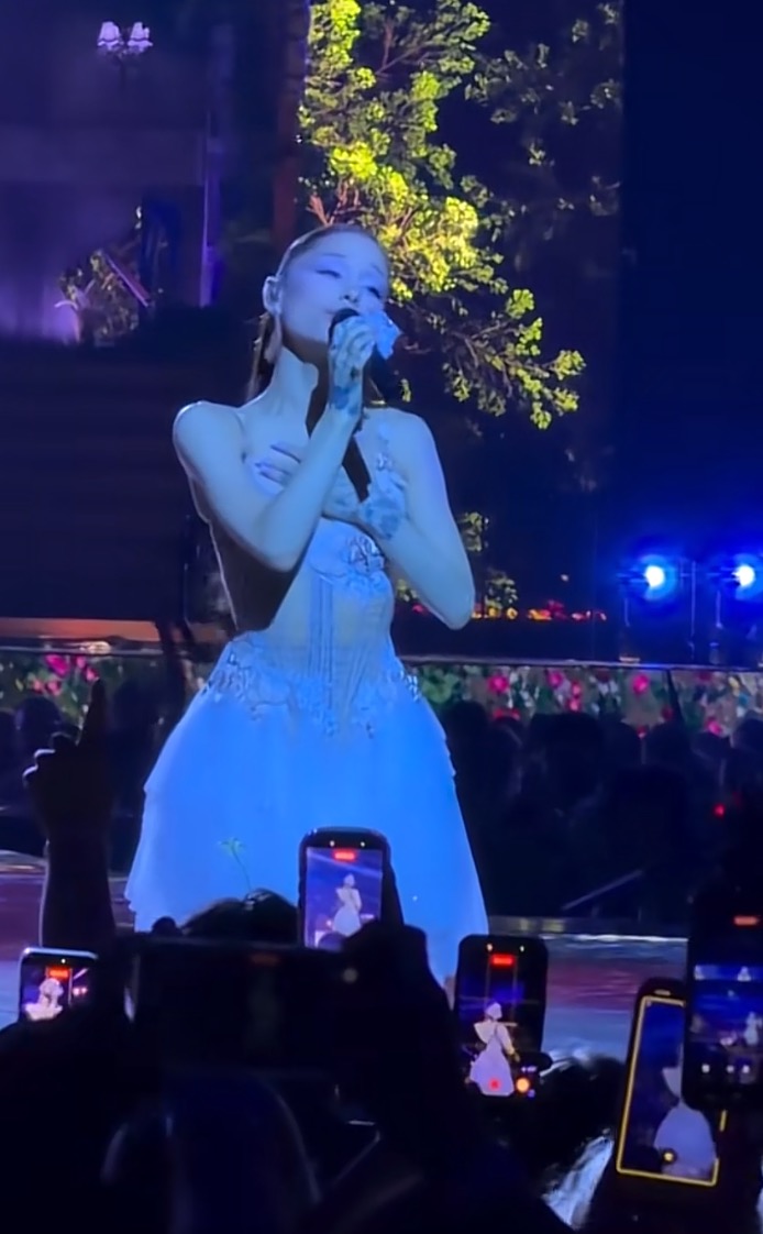 Ariana Grande singing on stage in a light blue gown with her hand on her chest.