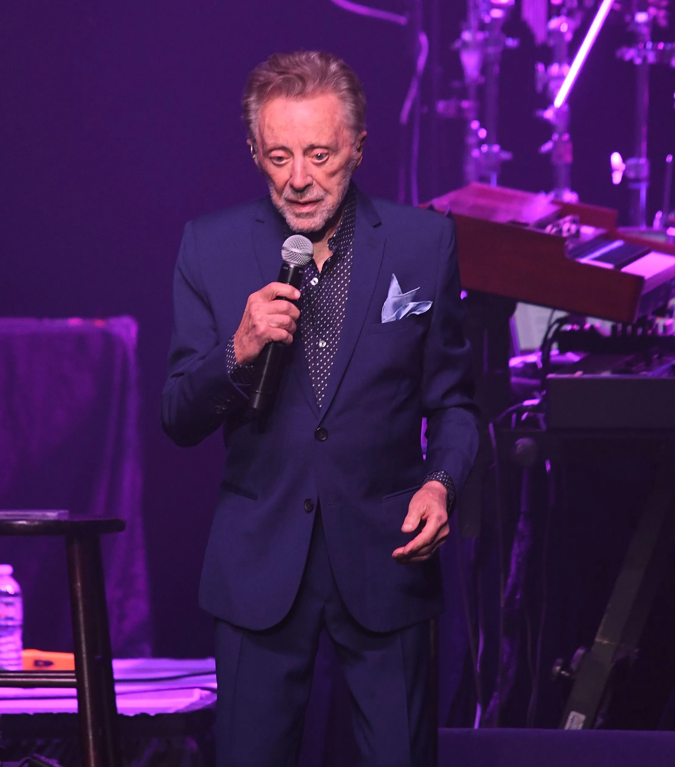 Frankie Valli performing on stage in a blue suit and patterned shirt, holding a microphone.